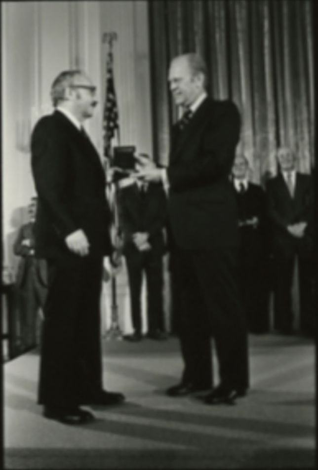 A black-and-white photograph of President Gerald Ford in a suit standing beside the seated mathematician George Dantzig, presenting him with a medal.