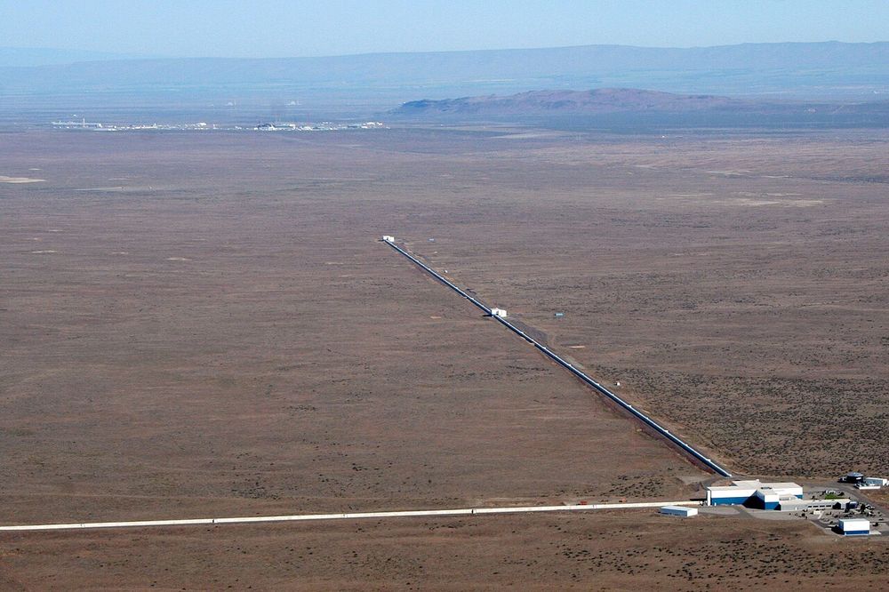 An aerial photograph of two long straight buildings meeting at a right angle in a flat desert landscape: the arms of the LIGO Hanford detector.