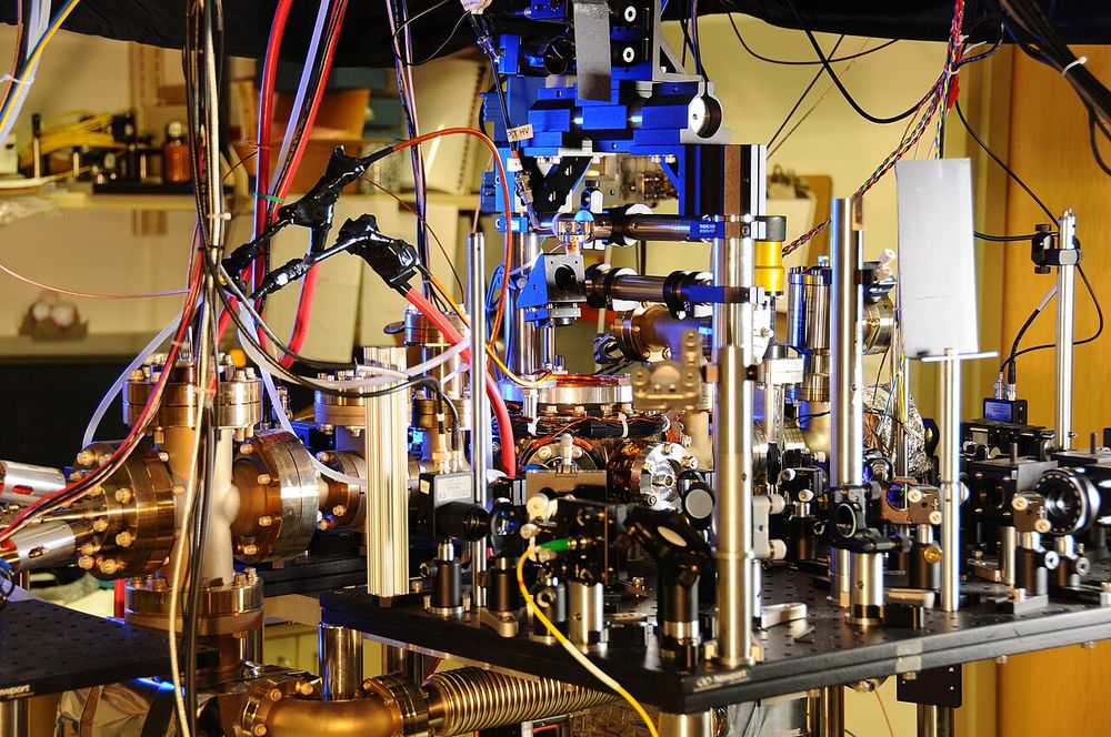 A dense laboratory apparatus of gold-coloured vacuum flanges, optical mounts, blue frames and cables around a central vacuum chamber.